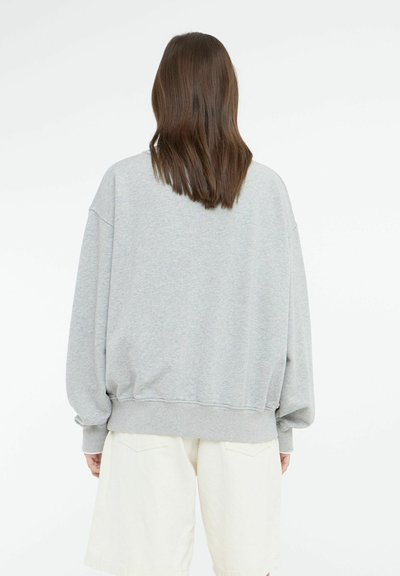 Felpa oversize grigia chiara con polsini e orlo a costine, caratterizzata da una texture morbida. Indossata con pantaloni corti chiari, vista da dietro.