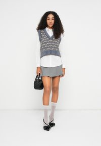 Mujer con cabello rizado vistiendo un chaleco de punto gris y azul con un patrón sobre una camisa blanca, falda gris plisada, calcetas blancas, tacones negros y sosteniendo un pequeño bolso negro.