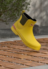 Botte de cheville en caoutchouc jaune avec un panneau latéral élastique noir et un accent rose sur la semelle. Texture lisse, design léger et bout arrondi.