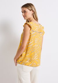 Top giallo con motivo a strisce zebra, maniche a sbuffo sfilacciate e dettagli in pizzo decorativi sul retro. Realizzato in tessuto morbido, design leggero.