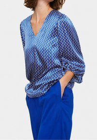 Donna che indossa una blusa blu a fantasia con scollatura a v e maniche a palloncino, abbinata a pantaloni blu, ambientata su uno sfondo bianco uniforme.