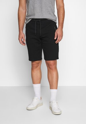 Mann, der schwarze Kordelzug-Shorts, weiße Socken, weiße Turnschuhe und ein graues T-Shirt trägt, steht vor einem weißen Hintergrund.