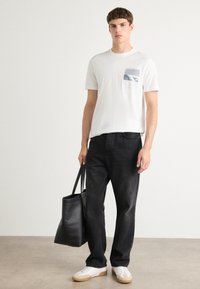 Hvid T-shirt med en lille blå grafisk lomme, sorte lige ben jeans, hvide sneakers med gummisåler og en sort tote bag.