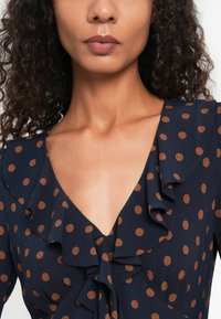 Marine jurk met bruine polka dots, voorzien van een gerimpelde halslijn en een diepe V-vorm, gemaakt van lichtgewicht stof. Gevoel van een textuur is duidelijk zichtbaar.