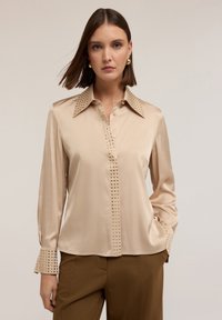 Beżowa bluzka satynowa z wyostrzonego kołnierza, ozdobiona kwadratowymi �ćwiekami wzdłuż przodu i mankietów, z długimi rękawami i prostym krojem.