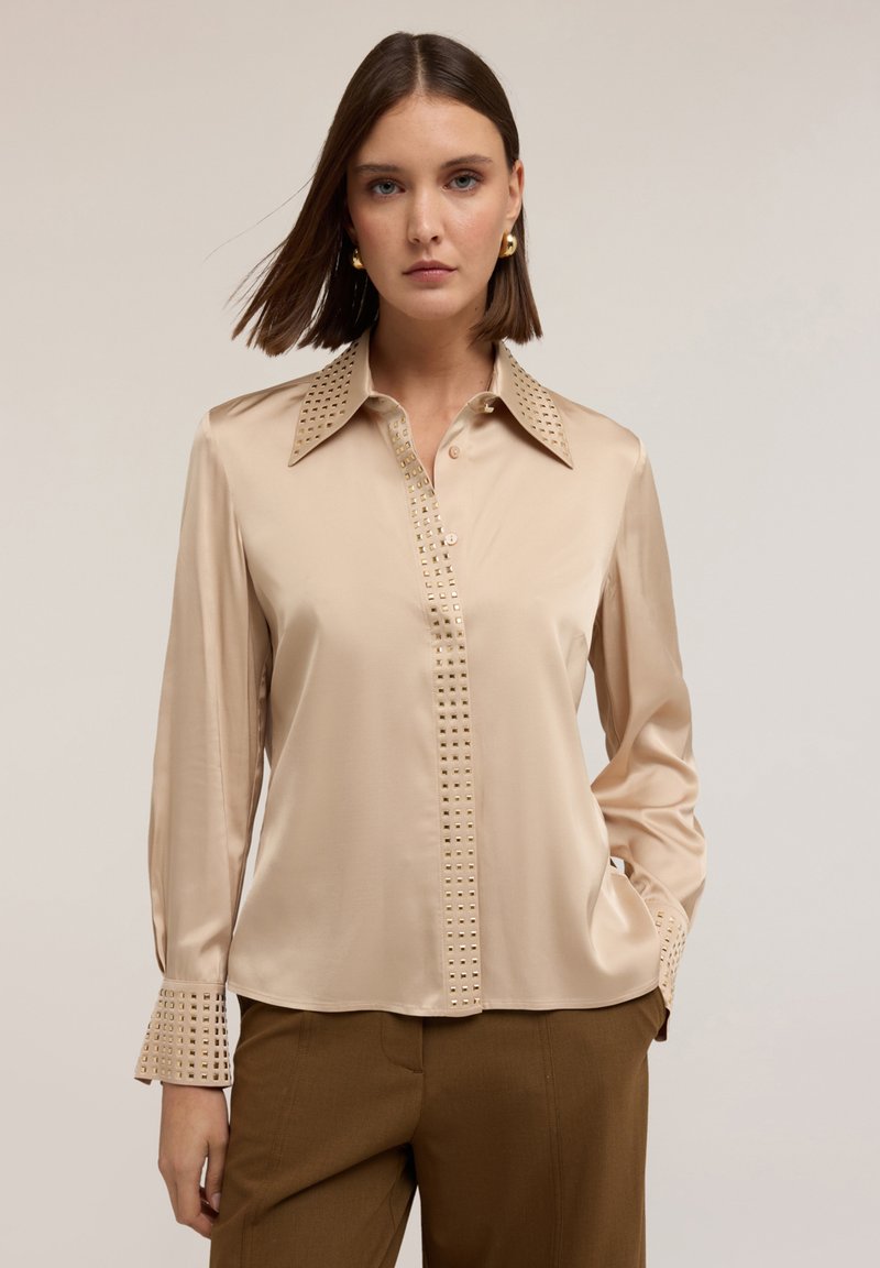 Beżowa bluzka satynowa z wyostrzonego kołnierza, ozdobiona kwadratowymi ćwiekami wzdłuż przodu i mankietów, z długimi rękawami i prostym krojem.