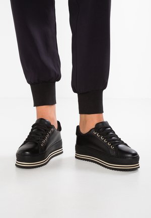 Personne portant des baskets compensées noires à lacets avec des semelles en caoutchouc rayées, associées à un pantalon de jogging noir retroussé aux chevilles.
