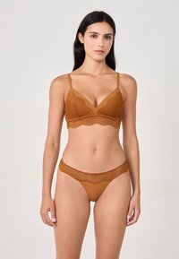 Donna con lunghi capelli neri che indossa un set di lingerie in pizzo marrone coordinato, in piedi contro uno sfondo grigio chiaro uniforme.