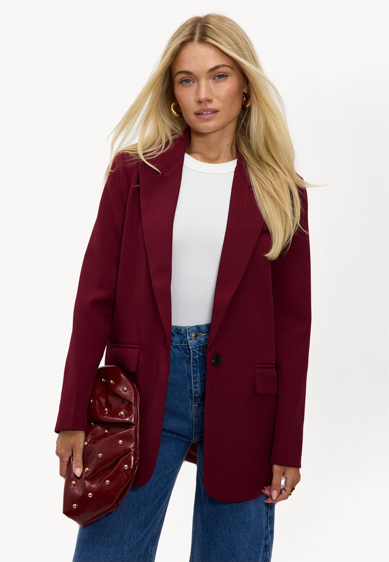 Bordeaux colbert met notch-revers, twee zakken en enkelsluiting; gecombineerd met een witte top en high-waisted blauwe jeans.