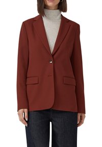Vrouw in een roestkleurige blazer met één knoop over een lichtgrijze coltrui en donkerblauwe jeans, staand tegen een witte achtergrond.