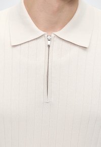 Gros plan d'une chemise en maille côtelée beige avec un col et une fermeture éclair argentée partiellement fermée au niveau de l'encolure.