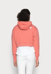 Cropt, met capuchon uitgevoerde sweatshirt in koraalkleur met een wit logo op de achterkant, gedragen over een wit shirt. Heeft lange mouwen en geribbelde manchetten.