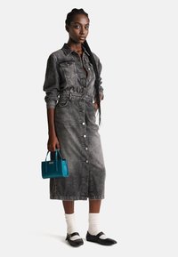 Abito midi in denim grigio sbiadito, con bottoni frontali, due tasche sul petto e vita con cintura. Abbinato a una borsa teal e ballerine nere.