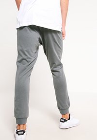 Person som bär grå joggingsbyxor och vita Nike-sneakers, stående med ryggen mot kameran mot en enkel ljus bakgrund.