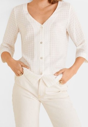 Femme portant un chemisier blanc boutonné à manches trois-quarts et un pantalon écru, les mains dans les poches avant.