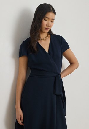 Kvinde med langt mørkt hår iført en marineblå omslagskjole med korte ærmer og en guldkædehalskæde, stående mod en ensfarvet lys væg.