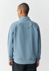 Mees lühikese juukselõikega, seljas helesinine fliisjope ribidega varrukaotste ja allääre ümber, seisab valge tausta ees näoga eemale.