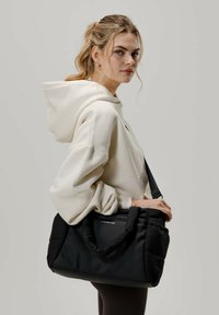 Musta pehmustettu kassi, jossa on kaksinkertaiset kahvat ja irrotettava olkahihna. Teksturoitu materiaali, jossa on tasainen paneeli ja logotiedot.