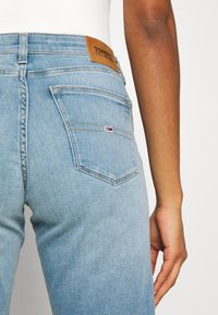 Jeans de mezclilla azul claro con una textura suave. Presentan un parche de cuero marrón etiquetado como "TOMMY JEANS" y un pequeño logotipo en rojo, blanco y azul.