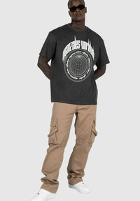 Dunkelgraues Grafik-T-Shirt mit einem runden Globusdesign in Weiß. Kombiniert mit beigen Cargo-Hosen mit mehreren Taschen und weißen Sneakern.