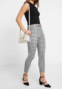 Donna che indossa un top senza maniche nero, pantaloni a quadri bianchi e neri, tacchi neri con cinturini alla caviglia e porta una borsa a tracolla bianca.