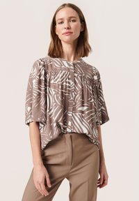 Bluză cu model maro și alb, cu mâneci scurte, închidere cu nasturi și detalii plisate; combinată cu pantaloni bej. Textură netedă.