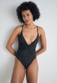 Μαύρο ολόσωμο μαγιό με βαθύ V-ντεκολτέ, στριμμένο μπροστινό σχέδιο, λεπτές τιράντες και υποτυπώδη υφή που γυαλίζει. Σχέδιο με ψηλή γραμμή ποδιού.