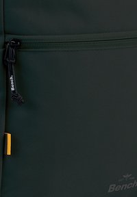 Mochila de tela verde con una superficie texturizada, cierre con cremallera y tirador negro, y una pestaña de acento amarilla. Incluye un detalle del logo de Bench.