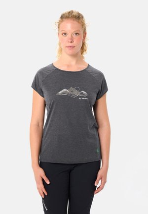 Femme aux cheveux blonds bouclés portant un t-shirt gris foncé imprimé de montagnes et un pantalon noir, debout devant un fond clair uni.