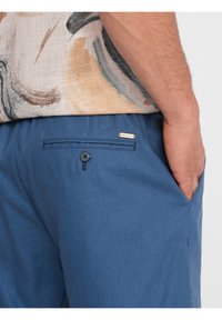 Shorts bleus en tissu léger, dotés d'une poche arrière avec une fermeture à bouton et d'un petit accent de marque. Texture lisse, design simple.