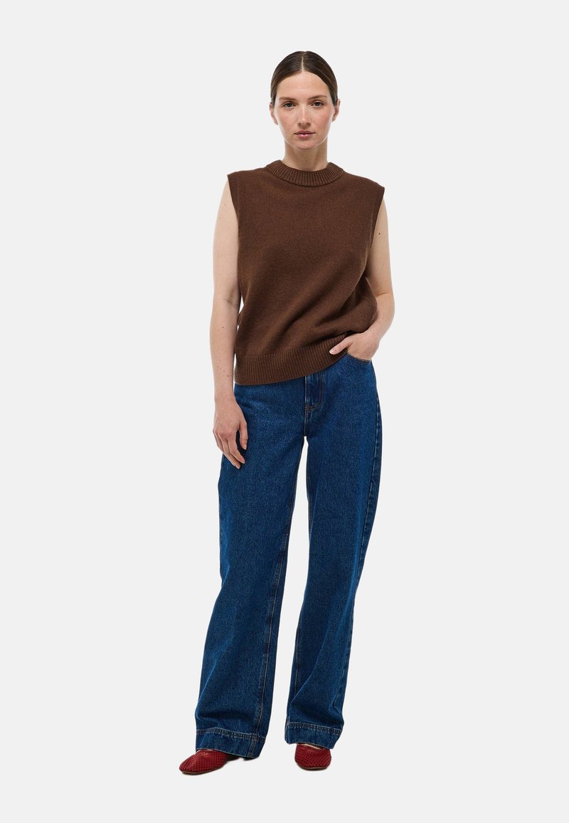 Débardeur en tricot marron sans manches avec un col montant côtelé, associé à un jean large bleu clair et des chaussures rouges à enfiler.