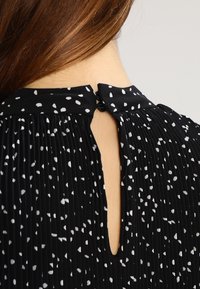 Blusa negra plisada con patrón salpicado blanco, que cuenta con una abertura en forma de llave en la parte posterior y un cierre de cinta en el cuello.