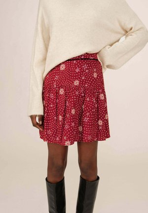 Jupe à motif floral rouge avec un design plissé, comportant une taille élastiquée et des accents décoratifs blancs et bleus. Accompagnée d'un pull crème.