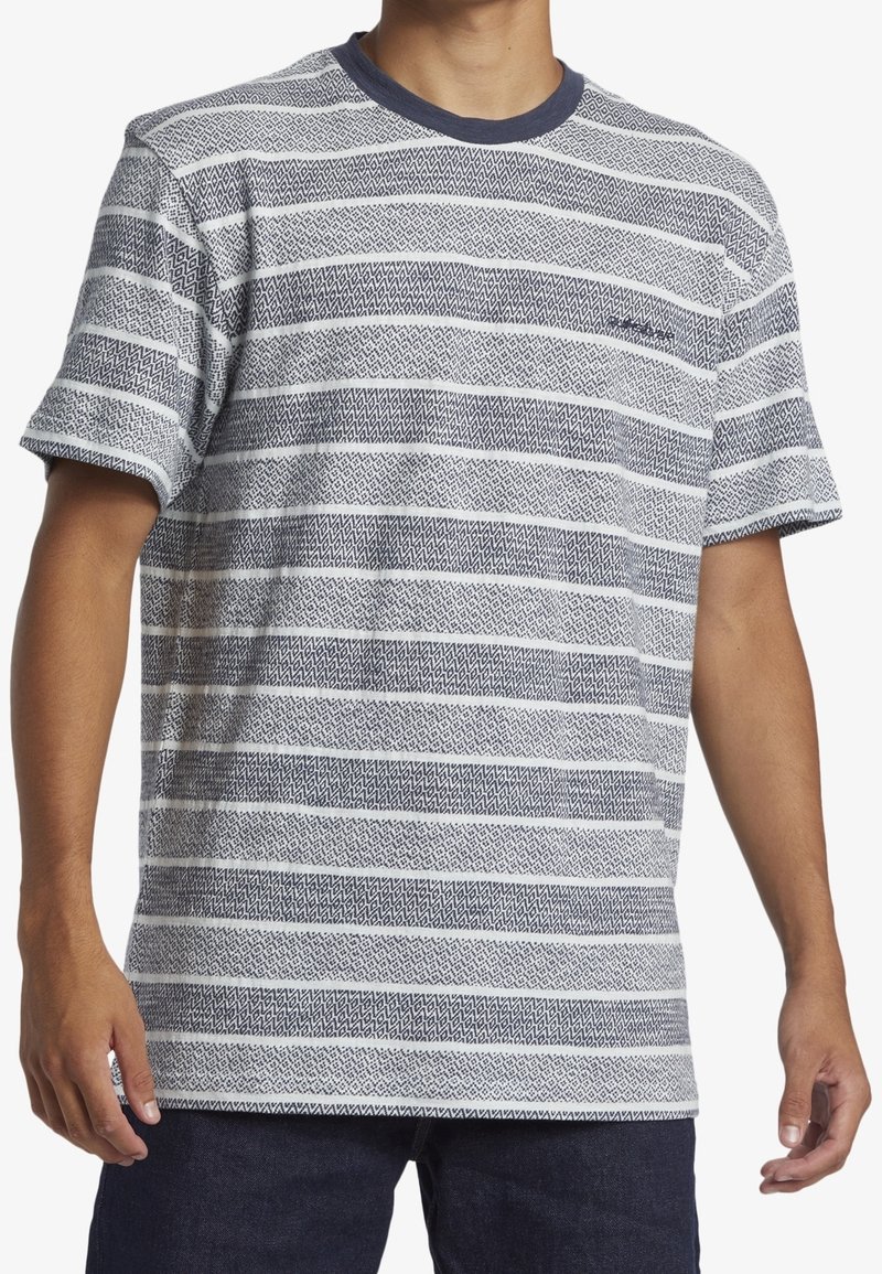 Homme portant un t-shirt à manches courtes gris et blanc rayé avec un col bleu foncé et un pantalon sombre.