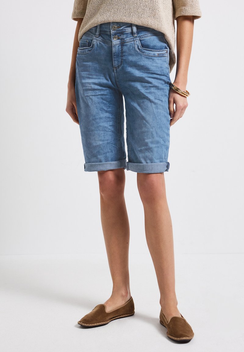 Bermudas de mezclilla azul claro con puños enrollados, con estilo de cinco bolsillos y cierre de botón. Se llevan con zapatos marrones de slip-on.