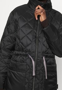 Chaqueta acolchada negra con un alto cuello, cierre de cremallera en la parte delantera, cinturilla elástica y dos bolsillos frontales. Presenta cordones de contraste y puños de punto.