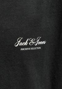 Μαύρο ύφασμα με λευκό κείμενο «Jack & Jones» σε καλλιγραφική γραφή και «ARCHIVE SELECTION» σε κεφαλαία, με γραμματοσειρά serif, τυπωμένα πάνω του.