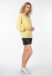 Gelber übergroßer Pullover mit langen Ballonärmel, kombiniert mit schwarzen Denim-Shorts und weißen Sneakern, vor einem schlichten Hintergrund.