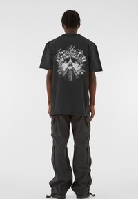 Mustblack graphic t-särk, mille tagaküljel on detailne disain, tekst "From Ashes Rise" ning keskne figuur, mida ümbritsevad lilleelemendid.