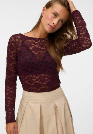 Femme portant un haut en dentelle florale bordeaux à manches longues et une jupe plissée beige, une main touchant ses cheveux.
