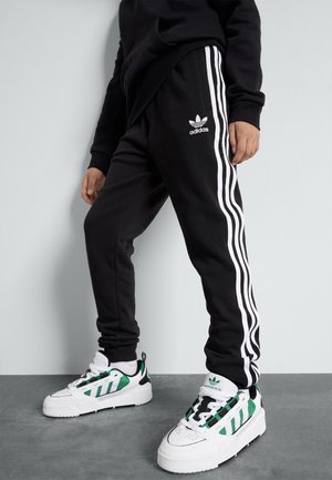 Personne portant un pantalon de survêtement Adidas noir avec des bandes blanches et des baskets blanches avec des détails verts et noirs, debout sur un sol gris.