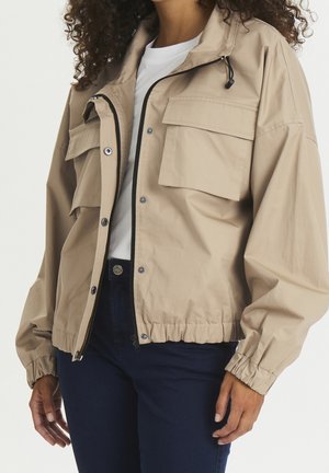 Kvinde iført en beige lynlåstrøje med store klaplommer over en hvid skjorte og mørkeblå bukser, stående i afslappet kropsholdning.