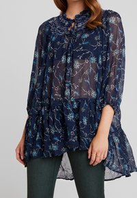 Námořnická květinová blůza s jemným krajkovým límcem, průhlednou tkaninou, dlouhými rukávy a vrstveným lemem. Obsahuje modré a bílé květinové vzory.