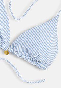 Haut de bikini rayé bleu et blanc avec fines bretelles et petit pendentif doré sur fond blanc.