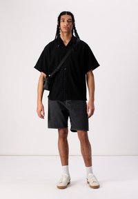 Camisa negra de manga corta y botonadura con un patrón texturizado, combinada con pantalones cortos de mezclilla oscuros. Calcetines blancos y zapatillas beige completan el conjunto.