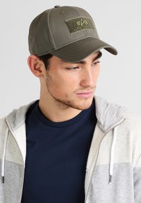 Oliven grøn baseballkasket med en flad skygge og et stofmærke mærket "ALPHA INDUSTRIES" på forsiden. Materiale i bomuldsblanding.