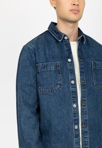 Donkere blauwe spijkerjas met knoopsluiting aan de voorkant, twee borstzakken en een klassieke kraag. De stof heeft een lichte textuur, typisch voor denim.