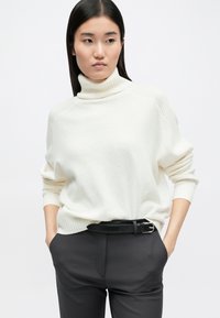 Λευκό πουλόβερ με λαιμόκοψη και ριγέ υφή, χαλαρή εφαρμογή και χαμηλούς ώμους, συνδυασμένο με σκούρες γκρι παντελόνες και μαύρη ζώνη.