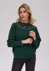 Zelen pulover iz pletene tkanine z teksturirano površino, z čipkastim vložkom okoli izreza. Model nosi črne kavbojke in zlat zapestnico.