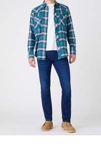 Groene en blauwe geruite flanellen shirt met twee borstzakken, gedragen over een wit T-shirt, gecombineerd met blauwe spijkerbroeken en tan suède schoenen.